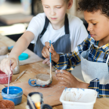 two children wearing aprons painting a tiny ceramic, handmade mug