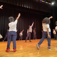 on a stage, a circle of kids follow along with a teacher in a dance class