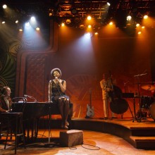African American woman in a black dress sitting on a piano while performing in Little Girl Blue: The Nina Simone Musical