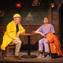 A man in a yellow raincoat and woman wearing purple sitting at a table onstage