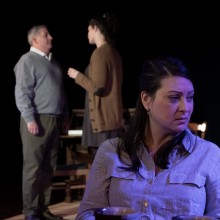 A scene onstage of a woman and a man talking and a woman listening to them sitting with a cup of coffee