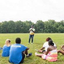 Storyteller standing before a group of people sitting on grass in a field.