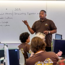 A man in a brown shirt is giving a lecture to students in front of a whiteboard.