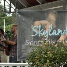 Two men standing on a porch playing guitar and singing behind a Skylands Songwriters Guild banner.