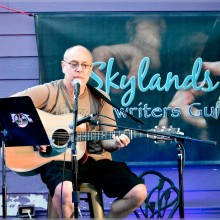 A man playing an acoustic guitar in  front of a sign that says Skylands Song writers Guild.