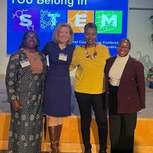 Four women pose for a photo in front of a sign that reads, "You Belong in STEM".