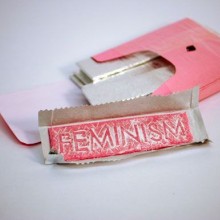 A pink box of gum with an unwrapped piece of gum the word feminism embossed into it.