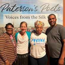 Four people pose for a photograph in front of a sign that reads Paterson's Poets.