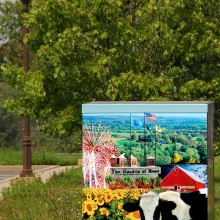 An outdoor mural  picture of a cow in a field of sunflowers with trees in the background.