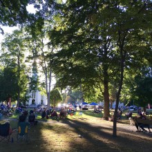 A wood glen with a white steeple church and people sitting on the grass and benches.