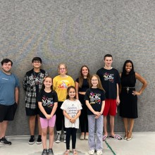 Students and teachers pose for a photo in front of a grey brick wall.