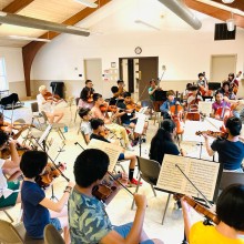 Children with string instruments in rehearsal in a bright studio classroom.