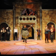 Students in period costumes performing on stage at the Walter K. Gordon Theater, under dramatic stage lighting.