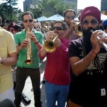 Trumpet and saxophone players wearing sunglasses, performing on a plaza for Make Music Day