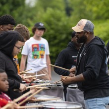 Young men engaged in drumming practice with an instructor providing guidance in an outdoor setting.