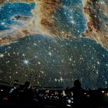 Musicians performing beneath a projected star field and nebula inside a planetarium, with audience seating visible in the fo