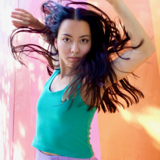A dancer with brown hair and arms overhead wearing a green tank top and lavender shorts with an orange and pink background