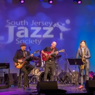 Six-piece jazz band performs onstage—keyboard, two guitars, drums, sax, and trumpet—with “South Jersey Jazz Society”