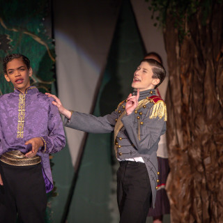 Two students perform a scene on stage in a school theater production, wearing period-style costumes and acting under stage l