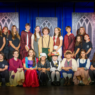 Full youth theater cast in period costumes poses on stage in front of a castle set, arranged in rows for a group photo.