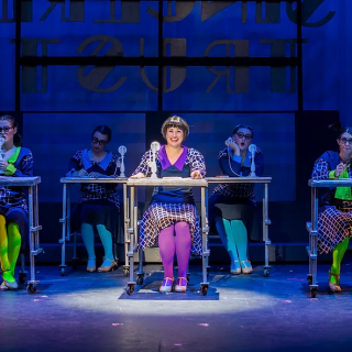 A group of people on a stage in 60s costumes at desks with spotlights on them