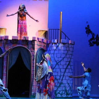 A woman stands on a balcony of a castle onstage singing, while men dance beneath her