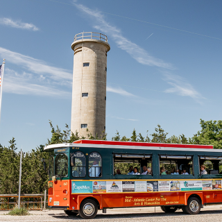 A vintage open-air green and orange tour bus drives by a lighthouse surrounded by pine trees