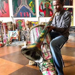 a man plays a spray painted bugle while drumming in a room of colorful paintings