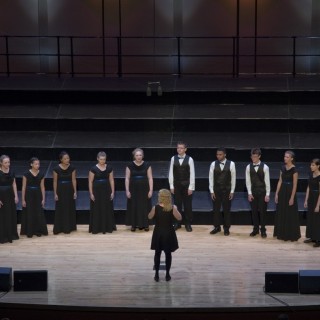 a semicircle of performers in black on a stage singing under the direction of a conductor