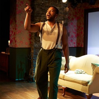 A man in suspenders standing in front of a couch is talking and pointing