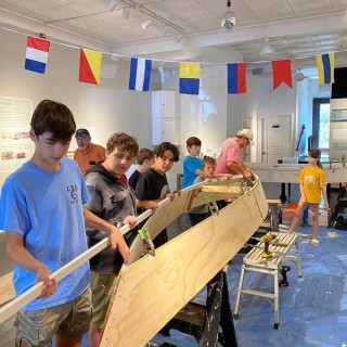 a line of preteen boys participate in building a wooden boat, with the help of a teacher, under a string of maritime flags