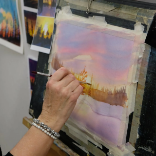 A hand paints a watercolor landscape of trees at sunset in the winter
