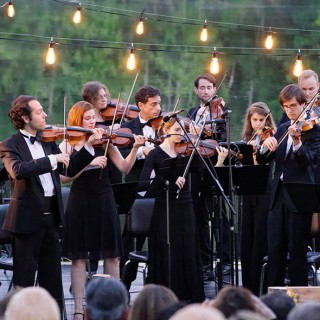 A group of people in tuxedos playing violins in front of a lake