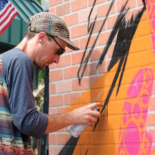 a man wearing sunglasses uses black spray paint to an outdoor mural