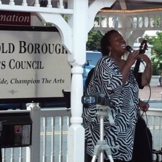 A woman singing in a white gazebo in front of a Freehold Borough Arts Council sign