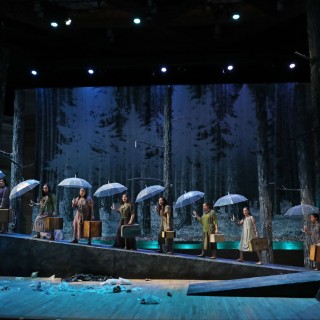 Actors on a dark and moody stage in a tableau holding umbrellas.