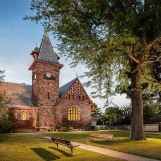 a large brown stone building from the 1800s with stained glass and clock tower beneath a large tree