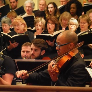 Photo of violinists performing in front of a singing choir.