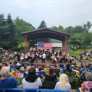 Rows of lawn chairs lined up in front of an orchestra band performing outside in a pavilion