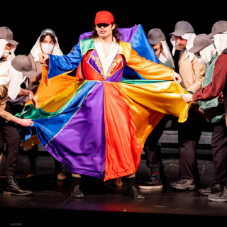 A group of people on a stage, one actor in the center dressed in a colorful coat