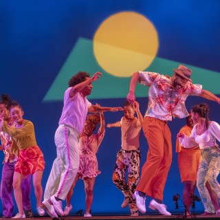 8 tap dancers in colorful outfits jumping in a cluster in front of a blue background with geometric sunset projection