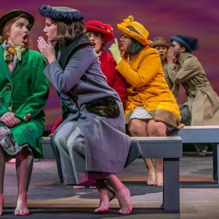 A cast of characters sitting on benches on stage whispering secrets.
