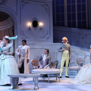 Seven actors on a white stage with a grand staircase wearing period clothing.