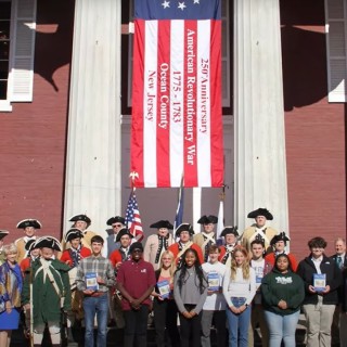 A group of people, Revolutionary War reenactors and teens, standing in front of a building with an American flag