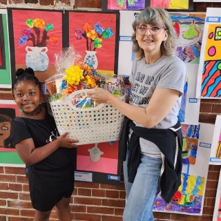 A woman holding a basket of art with a little girl in front of it.