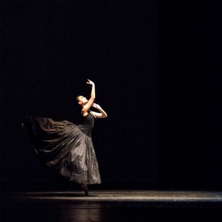 A ballerina in all black tutu poses on a darkened stage.