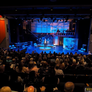 A theater stage with an audience of people seated in rows, attentively watching a performance.