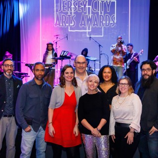 a multiracial group of 4 men and 4 women, standing in front of musicians on a stage at the 2022 Jersey City Art Awards cerem