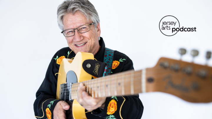 Richie Furay holding a guitar towards the camera with the Jersey Arts Podcast logo on top