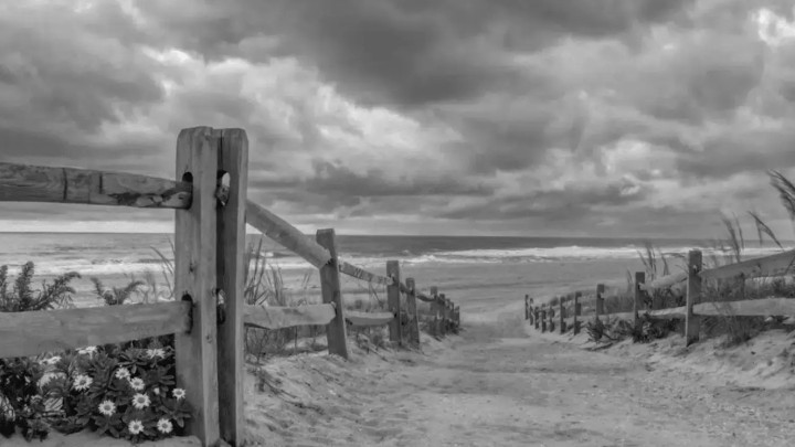 Photo of a beach walkway with a fence, grass, and a view of the ocean and clouds above
